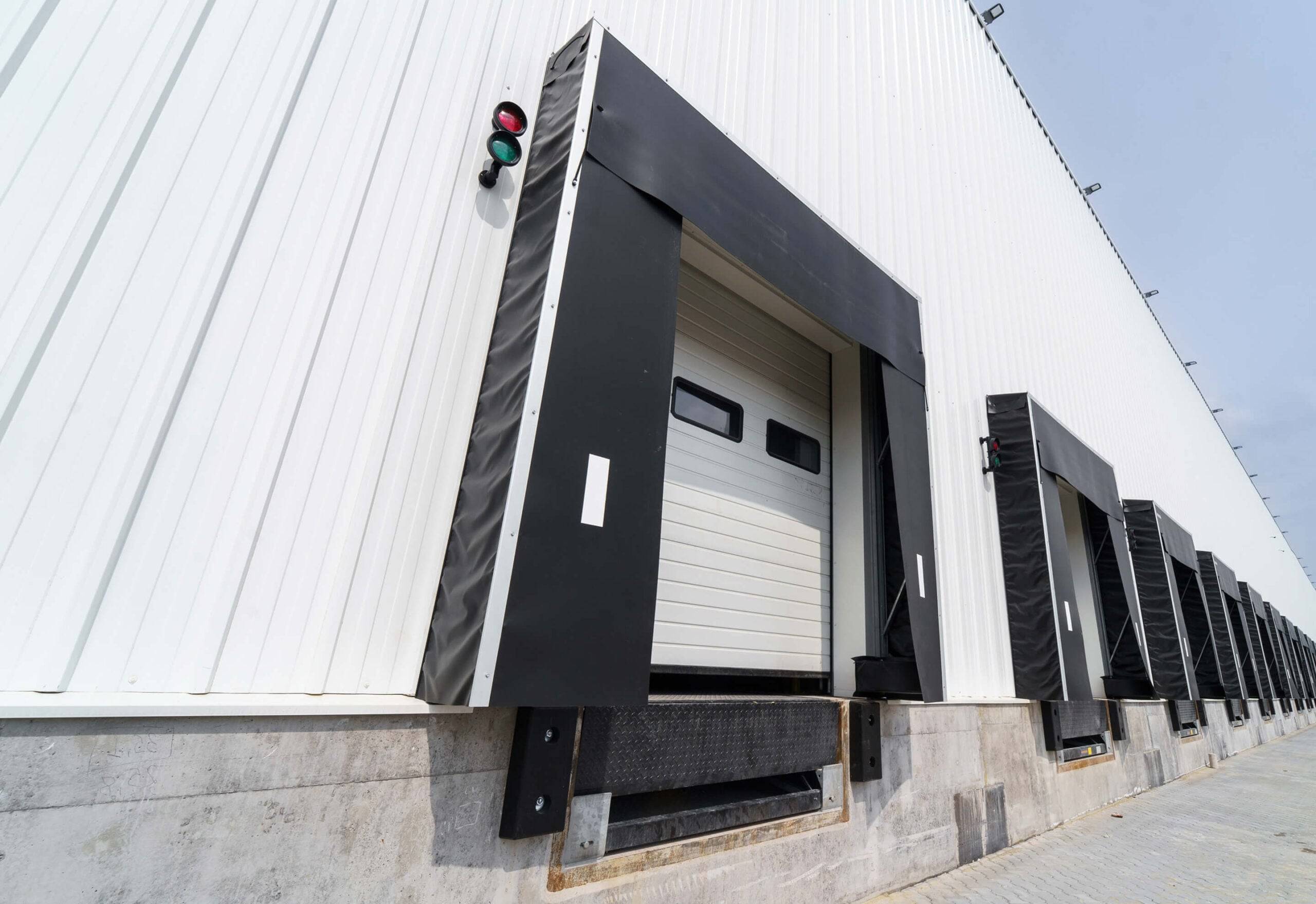 Multiple industrial loading dock doors with traffic lights mounted on black frames along a white warehouse