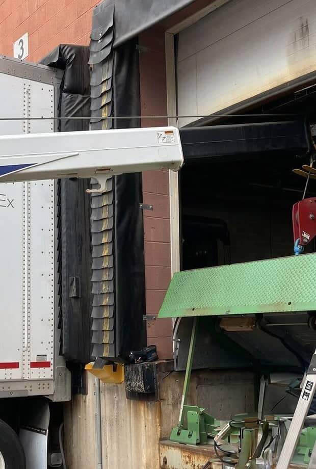 A loading dock with an open bay door showing a white truck with extended arm and black protective covering next to green